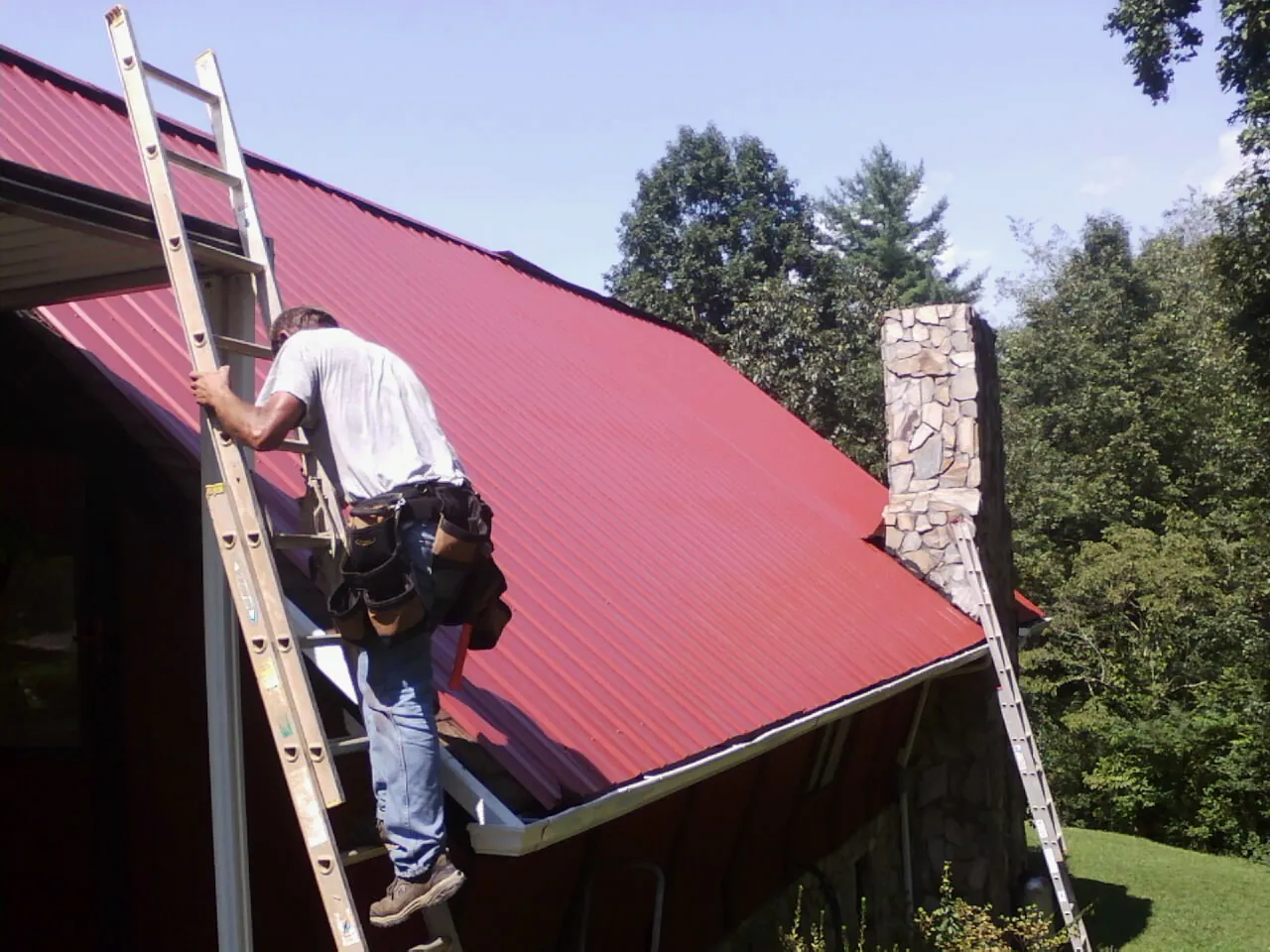 Roofer on a ladder beside a red metal roof and stone chimney — Soffit & Fascia Repair by Roofing Brothers in Uvalde
